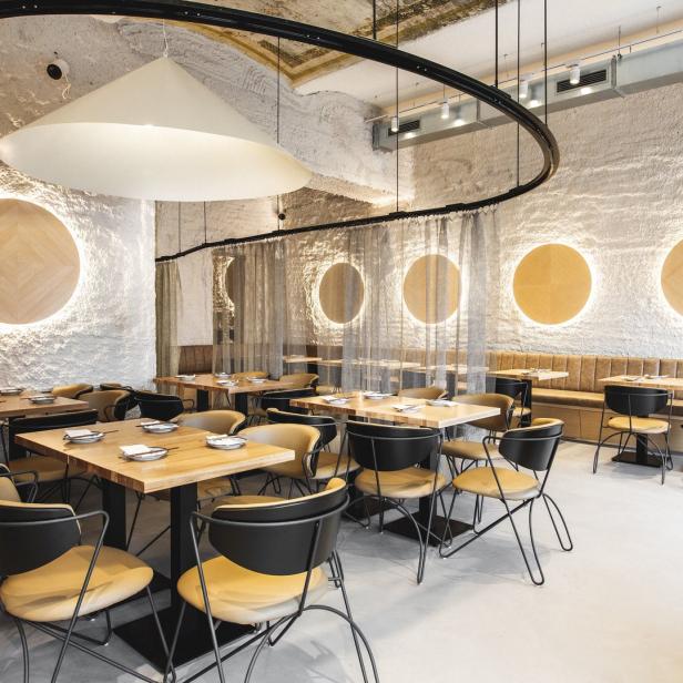 Innenansicht eines modernen Restaurants mit Holztischen und Stühlen vor einer strukturierten Wand mit kreisförmigen Leuchten.