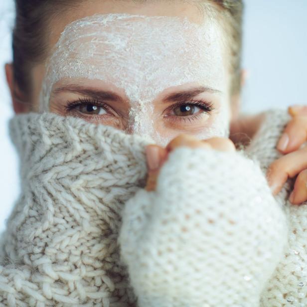 Eine Frau mit Gesichtsmaske kuschelt sich in einen dicken, weißen Strickpullover.