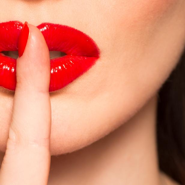Eine Frau mit rotem Lippenstift hält den Finger vor den Mund.