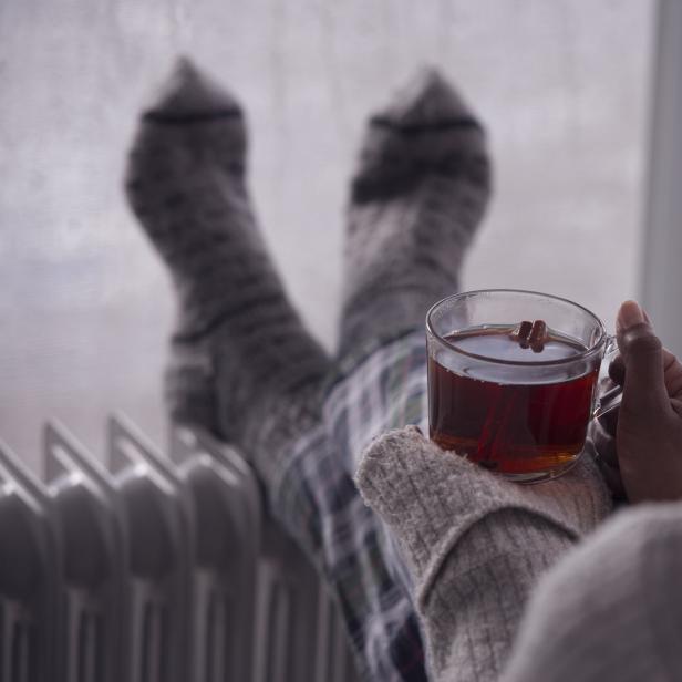 Eine Person mit Wollsocken wärmt sich mit einem Getränk vor einem Heizkörper.