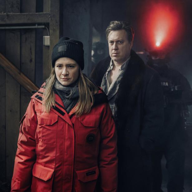 Eine Frau in roter Jacke und ein Mann stehen vor einem SWAT-Team mit rotem Licht.