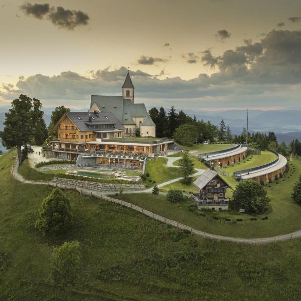 Ein Hotel und eine Kirche stehen auf einem grünen Hügel mit Blick auf eine Berglandschaft.
