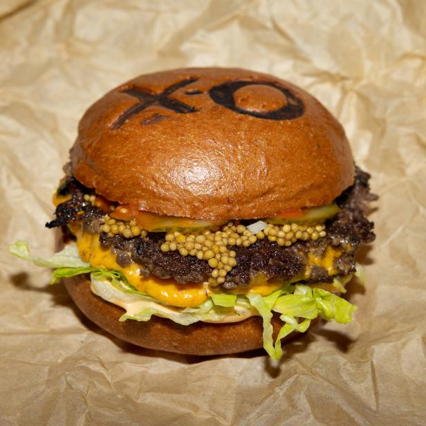 Ein doppelter Cheeseburger mit Salat, Gurken und Senf auf einem Brötchen mit „XO“-Markierung.