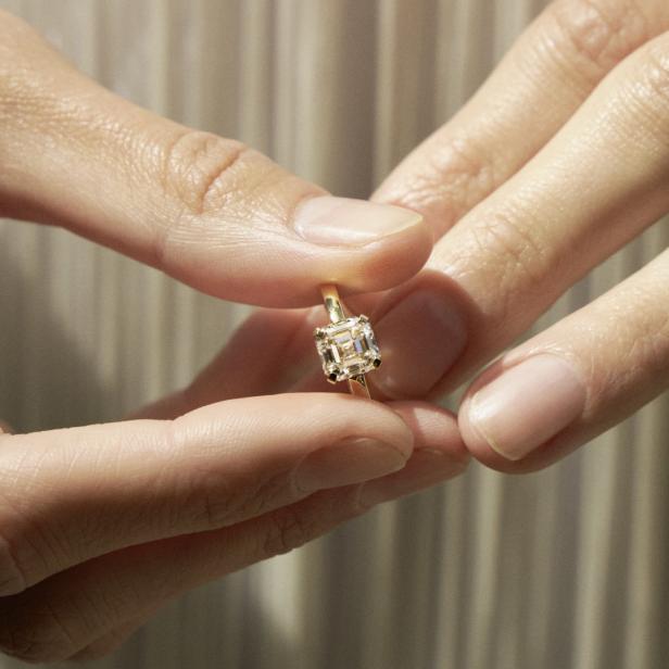 Ein goldener Verlobungsring mit einem quadratischen Diamanten wird von zwei Händen gehalten.