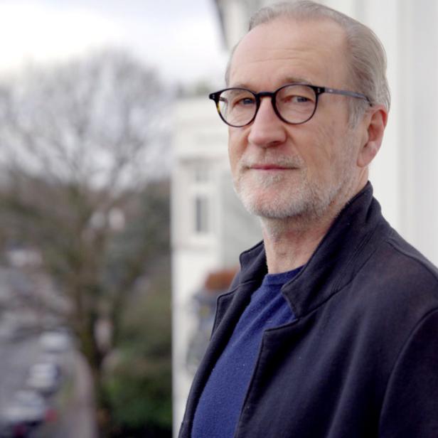 Ein Mann mit Brille steht auf einem Balkon und blickt in die Ferne.