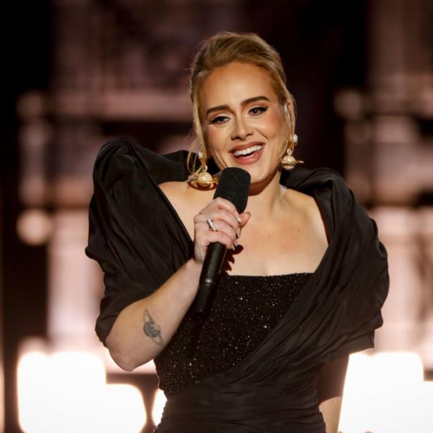 Adele singt in einem schwarzen Kleid mit glitzerndem Oberteil in ein Mikrofon.