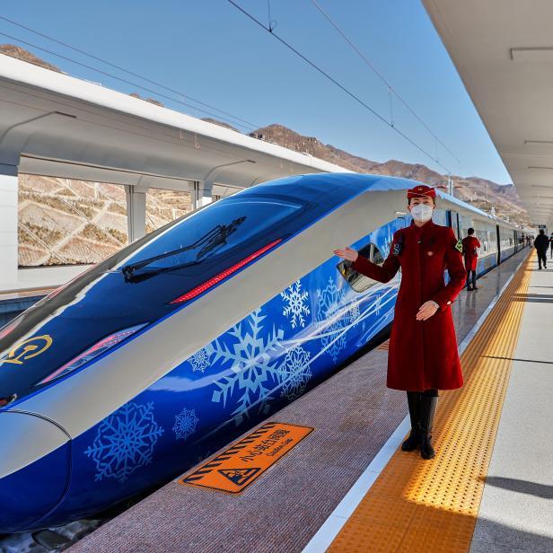 Eine Zugbegleiterin steht vor einem blau-weißem Hochgeschwindigkeitszug auf einem Bahnsteig.