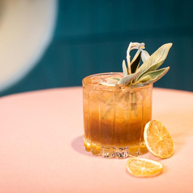 Ein Cocktail mit Eiswürfeln, Salbei und getrockneten Orangenscheiben auf einem Tisch.