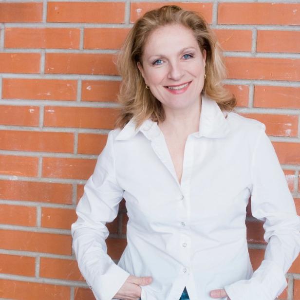 Eine Frau mit blonden Haaren lächelt vor einer roten Backsteinmauer.