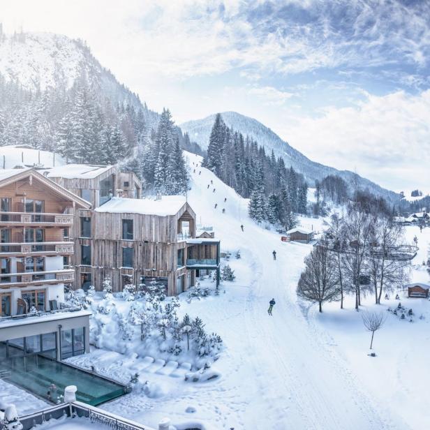 Winterlandschaft mit schneebedeckten Bergen, einem Hotel und Skifahrern auf der Piste.