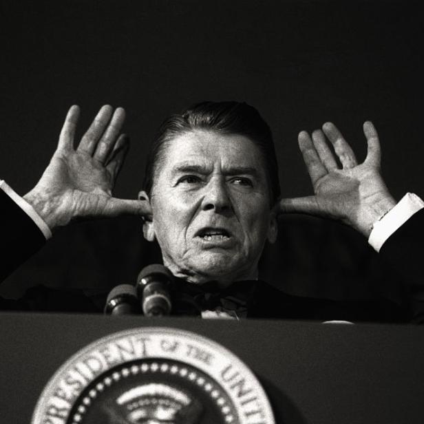 Ronald Reagan hält sich die Ohren zu, während er an einem Rednerpult steht.