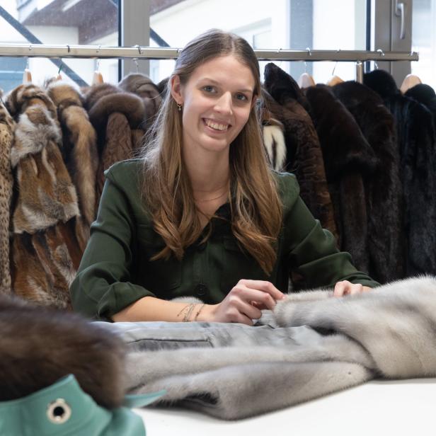 Eine Frau sitzt an einem Tisch mit einem Pelzmantel, im Hintergrund hängen weitere Pelze.