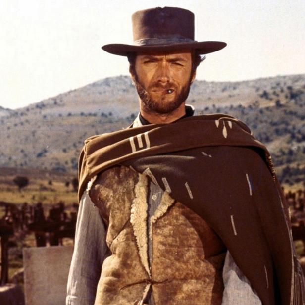 Clint Eastwood als „Blondie“ vor einer Hügellandschaft mit einem Friedhof im Hintergrund.