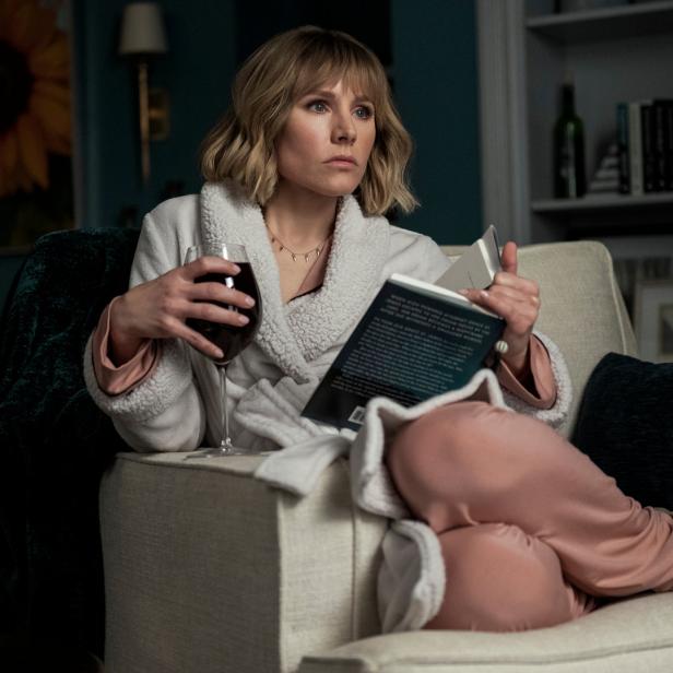 Eine Frau im Bademantel sitzt mit einem Buch und einem Glas Wein auf einem Sofa.