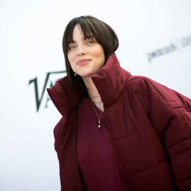 Billie Eilish trägt eine weinrote Jacke und steht vor einem weißen Hintergrund.