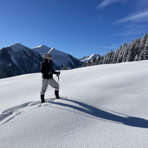 Eine Frau steht mit Wanderstöcken im Schnee, im Hintergrund sind Berge und Bäume zu sehen.