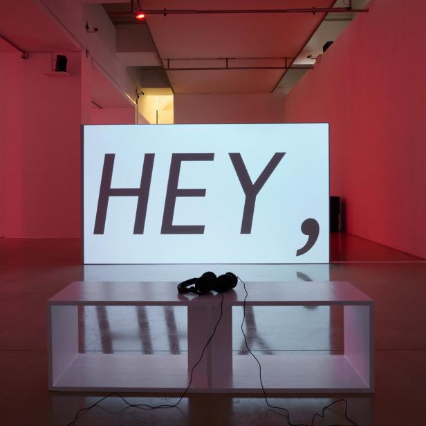 In einem Ausstellungsraum steht eine Leinwand mit dem Wort „HEY,“ darauf. Davor liegen Kopfhörer auf einem weißen Podest.