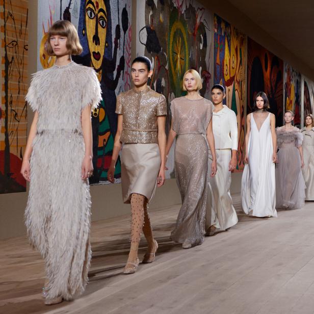 Models präsentieren elegante Kleider auf einem Laufsteg vor farbenfrohen Wandteppichen.