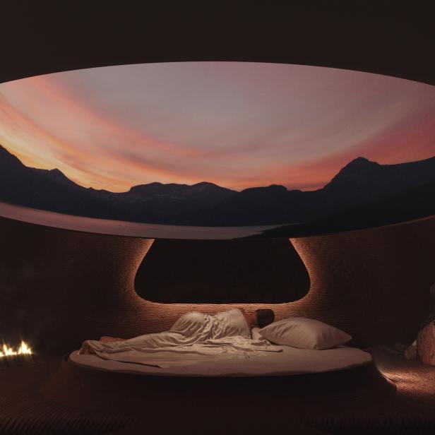 Eine Person schläft in einem runden Bett in einem Raum mit Blick auf eine Berglandschaft im Sonnenuntergang.