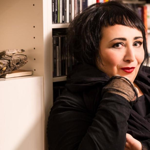 Eine Frau mit dunklen Haaren posiert vor einem Bücherregal mit einer Echse.