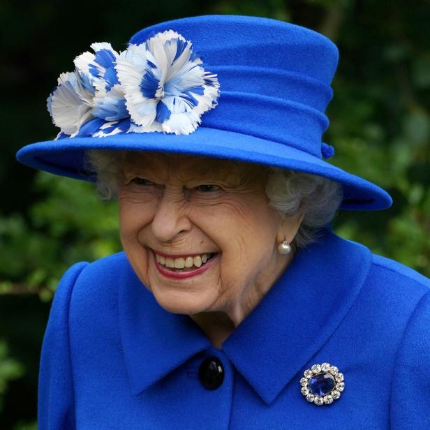 Königin Elisabeth II. lächelt in einem blauen Hut mit Blumenapplikation.