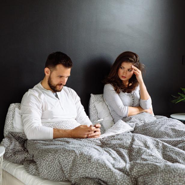 Ein Mann tippt im Bett auf seinem Handy, während eine Frau besorgt daneben sitzt.