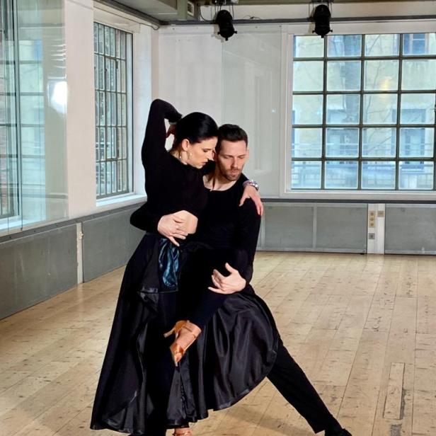 Ein Tanzpaar in schwarzer Kleidung tanzt in einem hellen Raum.