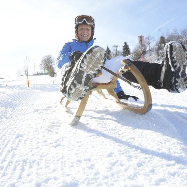 Eine Frau mit Helm und Skibrille fährt auf einer Holzrodel den Hang hinunter.