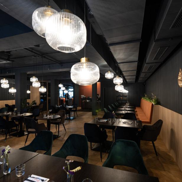 Innenansicht eines eleganten Restaurants mit dunklen Tischen und Stühlen unter dekorativen Lampen.