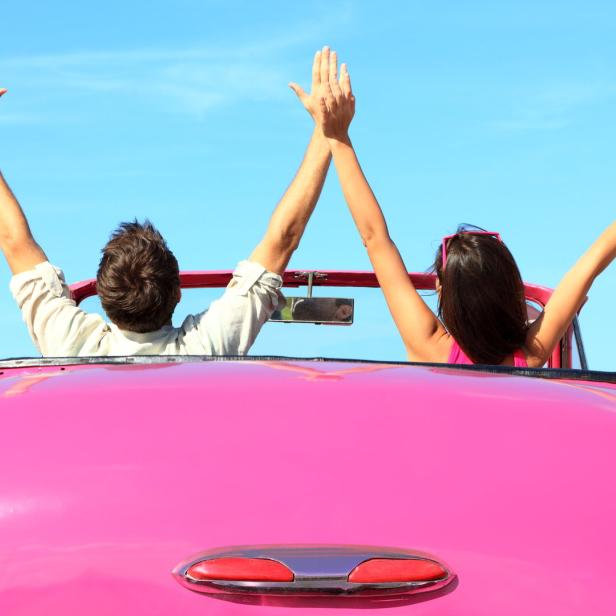 Ein Paar fährt mit einem pinkfarbenen Cabriolet und reckt die Arme in die Luft.