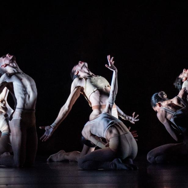 Fünf Tänzerinnen und Tänzer in beigen Kostümen führen eine synchrone Choreografie auf einer dunklen Bühne auf.