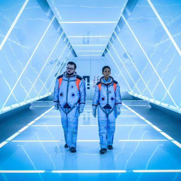 Zwei Astronauten gehen in Raumanzügen einen hell erleuchteten, futuristischen Korridor entlang.
