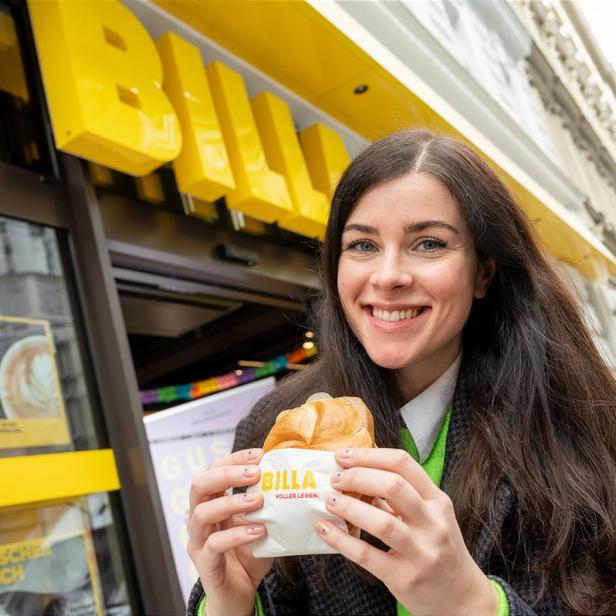Eine lächelnde Frau hält ein belegtes Brötchen vor einem Billa-Supermarkt.