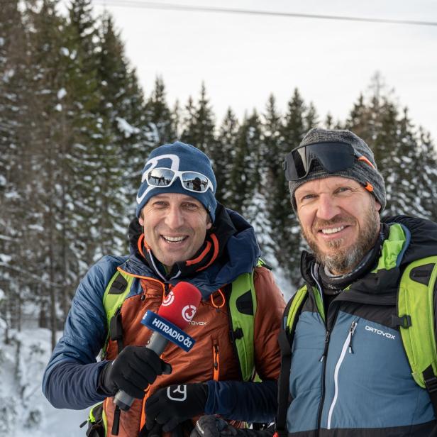 Zwei Männer stehen im Schnee vor einem Wald, einer hält ein Mikrofon von Hitradio Ö3.
