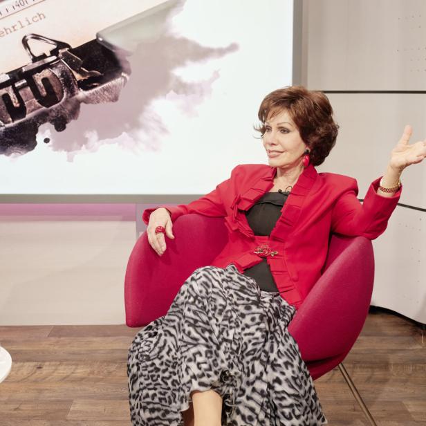 Eine Frau in roter Jacke sitzt in einem Sessel vor einem Bildschirm mit Schreibmaschinenmotiv.