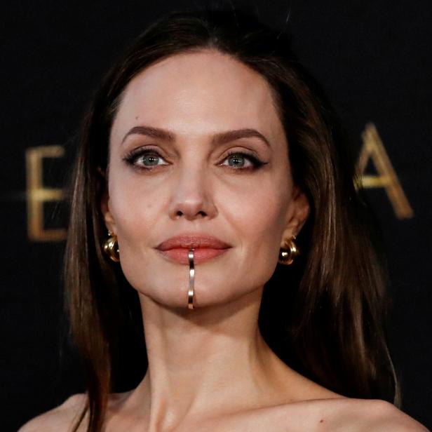 Angelina Jolie mit einem goldenen Schmuckstück unterhalb ihrer Lippe vor dem Hintergrund des Filmtitels „Eternals“.