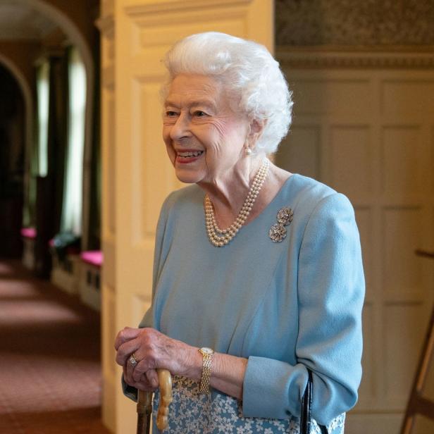 Königin Elizabeth II. lächelt mit einem Gehstock in einem Korridor.