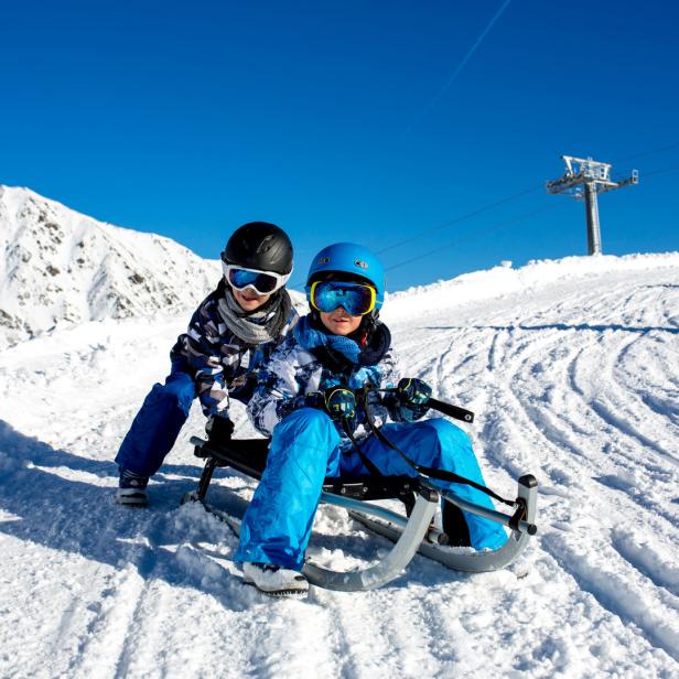 Zwei Kinder fahren mit einem Rodelschlitten auf einer Skipiste.