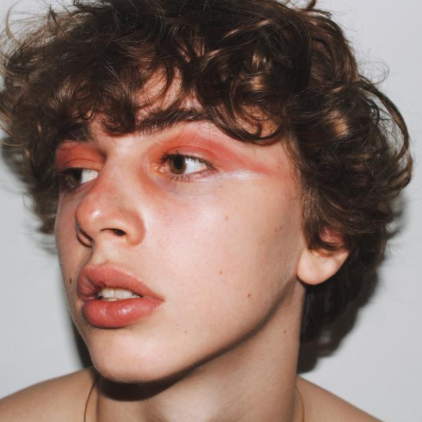 Ein junger Mann mit lockigem Haar und auffälligem, orangefarbenem Make-up blickt zur Seite.