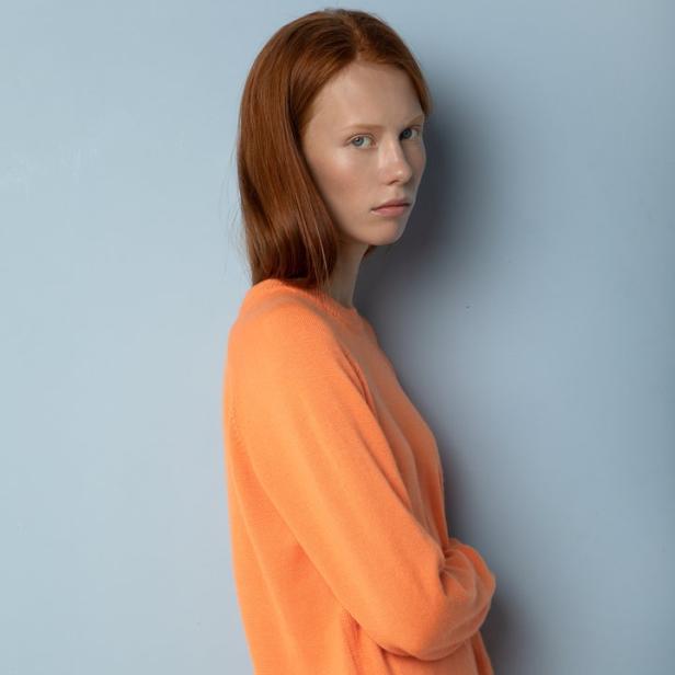 Eine Frau mit roten Haaren trägt einen orangefarbenen Pullover und eine rote Hose.