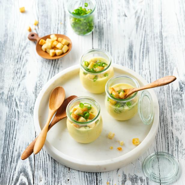 Drei Gläser mit gelber Suppe, Croutons und Schnittlauch auf einem Tablett.