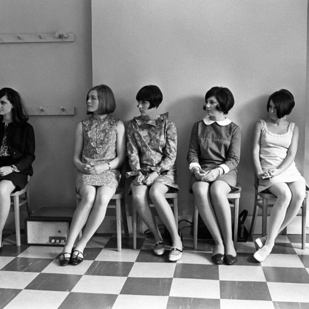 Sechs junge Frauen im Stil der 1960er Jahre sitzen auf Stühlen vor einer Wand.