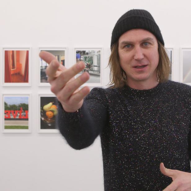 Ein Mann mit Mütze gestikuliert vor einer Wand mit gerahmten Fotos.