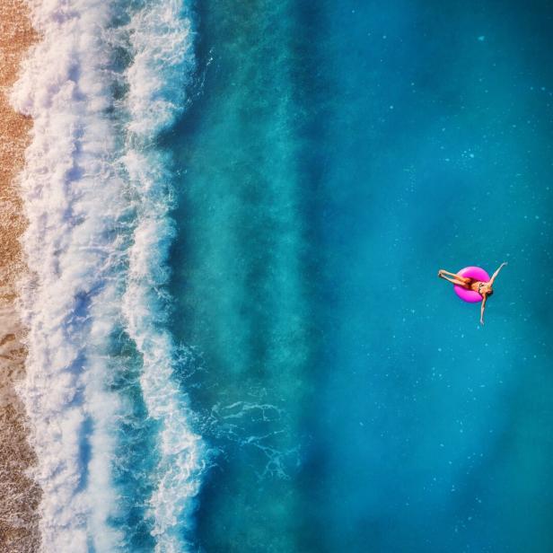 Aerial view of young woman swimming on the pink swim ring in the transparent turquoise sea in Oludeniz. Summer seascape with girl, beach, beautiful waves, blue water at sunset. Top view from drone