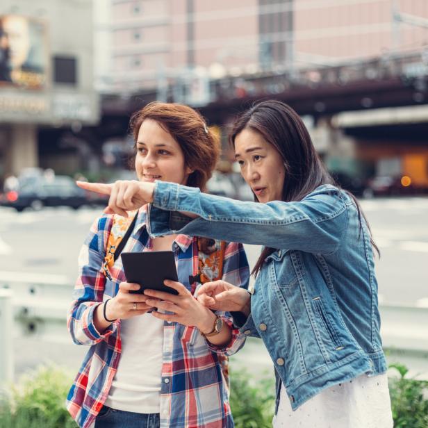 Zwei Frauen stehen in einer Stadt und schauen auf ein Tablet, während eine von ihnen in die Ferne zeigt.