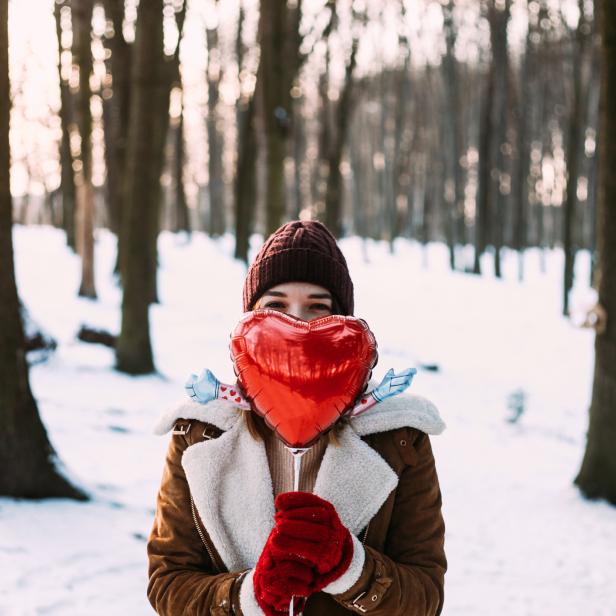 Eine Frau mit roter Mütze hält einen herzförmigen Ballon vor dem Gesicht in einem verschneiten Wald.