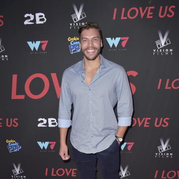Ein Mann posiert auf einem roten Teppich vor einer Wand mit Logos für den Film „I Love Us“.