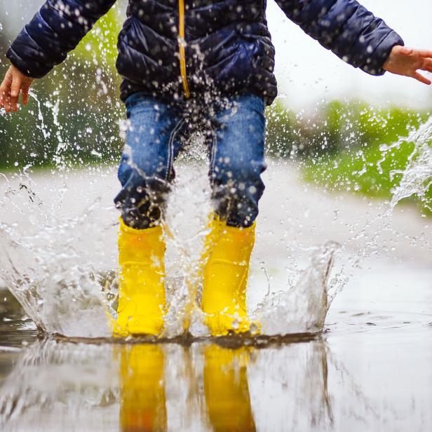 Ein Kind mit gelben Gummistiefeln springt in eine Pfütze, wodurch das Wasser hochspritzt.