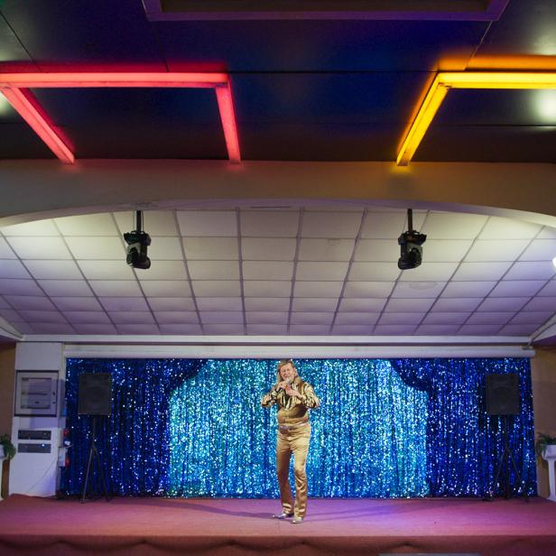 Ein Mann in einem goldenen Anzug steht auf einer Bühne mit blauem Glitzerhintergrund.
