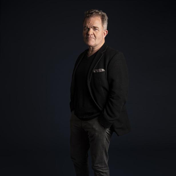 Ein Mann in Jeans, Sakko und braunen Lederschuhen steht vor einem dunklen Hintergrund.